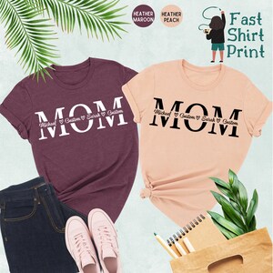 Puede incluir: Dos camisetas con la palabra "MOM" impresa en letras grandes. Las camisetas son de color granate jaspeado y melocot&oacute;n jaspeado. La palabra "MOM" est&aacute; rodeada de peque&ntilde;os corazones y nombres, incluyendo "Michael", "Custom", "Sarah" y "Custom".