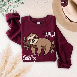 K&ouml;nnte beinhalten: Burgunderfarbenes Sweatshirt mit einem Cartoon-Faultier auf einem Ast, mit dem Text "A SLOTH does more" und "work than my PANCREAS". Das Faultier ist braun und l&auml;chelt. Das Sweatshirt ist gefaltet.