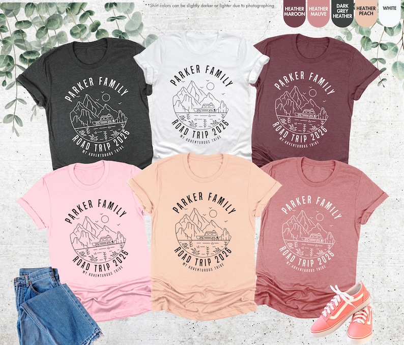 May include: A selection of short-sleeved t-shirts in shades like grey, white, pink, peach, and maroon. Each shirt displays a circular graphic with the text "PARKER FAMILY ROAD TRIP 2026" and a mountain and camper illustration. A pair of pink trainers and jeans are also visible.
