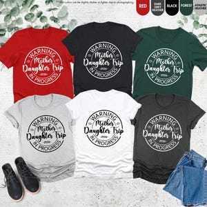 K&ouml;nnte beinhalten: Eine Auswahl an T-Shirts in verschiedenen Farben, darunter Rot, Schwarz und Gr&uuml;n. Jedes Shirt zeigt eine runde Grafik mit dem Text "WARNING Mother Daughter Trip -2026- IN PROGRESS" in Wei&szlig;. Die Shirts werden mit einem Paar schwarzer Stiefel und einer Jeans pr&auml;sentiert.