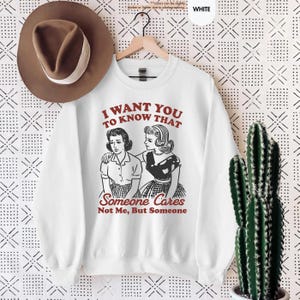 May include: White sweatshirt with the text "I WANT YOU TO KNOW THAT Someone Cares Not Me, But Someone" in red. The graphic features a vintage illustration of two women. A brown hat hangs above the sweatshirt.