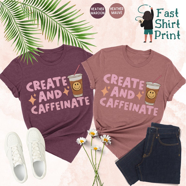 May include: Two t-shirts with the text "Create and Caffeinate" in pink letters, a coffee cup with a smiley face, and pink stars. The shirts are heather maroon and heather mauve.