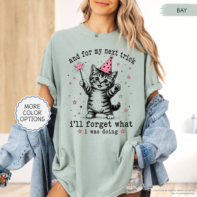 May include: A sage green t-shirt featuring a black and white illustration of a kitten wearing a pink party hat and holding a star-tipped wand. The text reads, and for my next trick i'll forget what i was doing, surrounded by small pink stars.