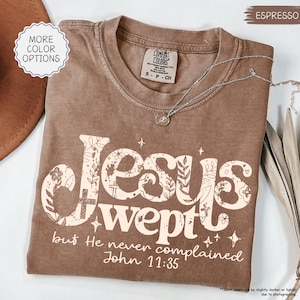 May include: Espresso-colored t-shirt with the words "Jesus wept" in a floral design. The text "but He never complained John 11:35" is printed below. A silver necklace with a pendant is draped on the shirt.