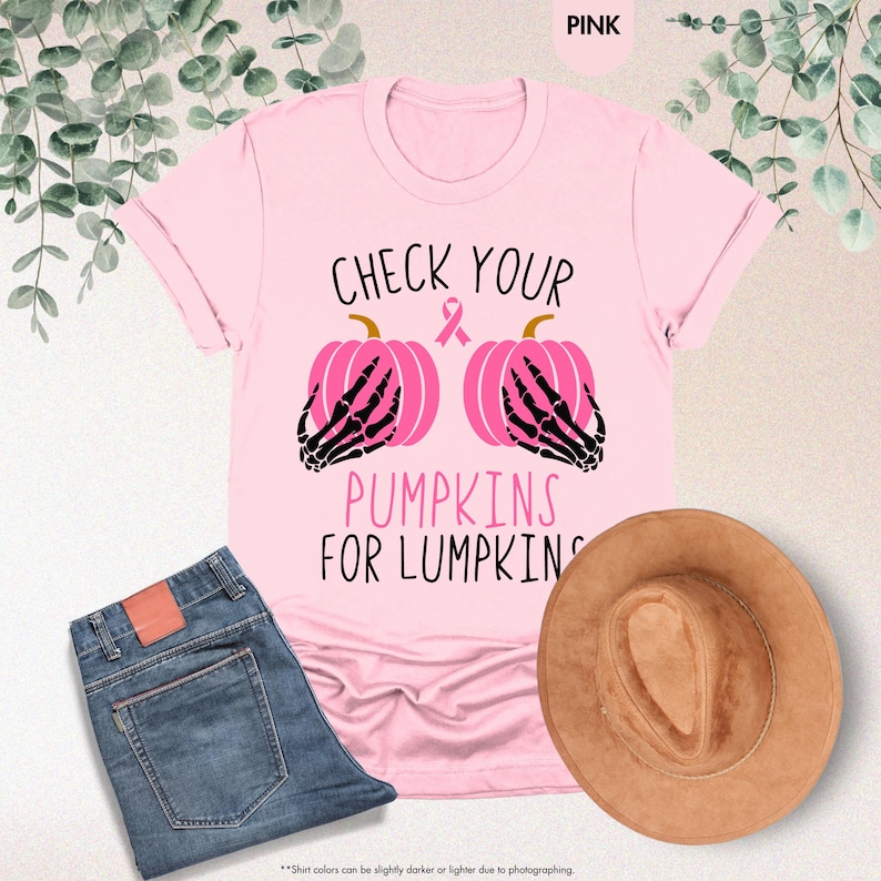 以下が含まれることがあります： 「CHECK YOUR PUMPKINS FOR LUMPKINS」の文字が入ったピンク色のTシャツ。2つのピンク色のカボチャと黒い骸骨の手、ピンクのリボンが描かれています。青いジーンズと茶色の帽子も画像に写っています。