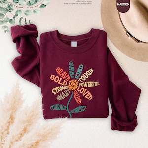 Pode incluir: Camisola bord&ocirc; com um design floral com palavras como "Corajoso", "Forte" e "Gentil". A parte superior de manga comprida tem gola redonda e punhos enrolados. O texto "You are enough" est&aacute; no centro do design floral.