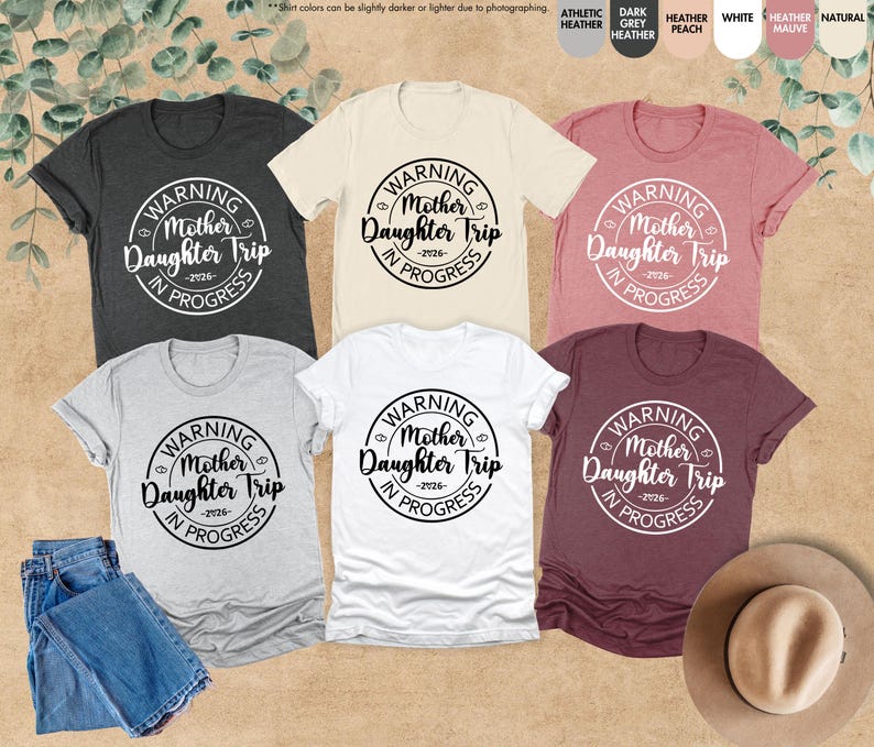 K&ouml;nnte beinhalten: Eine Auswahl an kurz&auml;rmeligen T-Shirts in verschiedenen Farben, darunter Grau, Wei&szlig; und Burgunderrot. Jedes Shirt zeigt eine runde Grafik mit dem Text "WARNING Mother Daughter Trip -2026- IN PROGRESS".