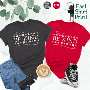 K&ouml;nnte beinhalten: Zwei T-Shirts mit dem Text "Be Kind" in wei&szlig;en Buchstaben, umgeben von kleinen wei&szlig;en Blumen. Die Shirts sind in dunkelgrau meliert und rot.
