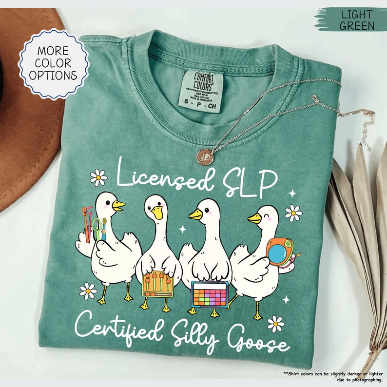 May include: Light green t-shirt with the text "Licensed SLP Certified Silly Goose" and a graphic of four white geese holding speech therapy tools. A silver necklace with a pendant is on the shirt.