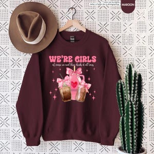 K&ouml;nnte beinhalten: Ein maroonfarbener Pullover mit dem Text "WE'RE GIRLS of course we need three drinks at all times" in Pink. Die Grafik zeigt drei rosa Getr&auml;nke mit rosa Schleifen und einem Herzen.