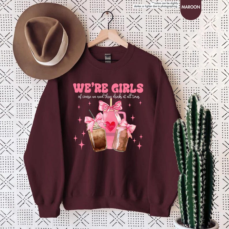 K&ouml;nnte beinhalten: Ein maroonfarbener Pullover mit dem Text "WE'RE GIRLS of course we need three drinks at all times" in Pink. Die Grafik zeigt drei rosa Getr&auml;nke mit rosa Schleifen und einem Herzen.