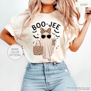 以下が含まれることがあります： 「BOO-JEE」の文字がプリントされたアイボリー色のTシャツ。サングラスとヒョウ柄のリボンをつけたお化けが、ヒョウ柄のトートバッグとコーヒーカップを持っています。黒いコウモリと星がお化けを囲んでいます。「MORE COLOR OPTIONS」は青い円の中にあります。