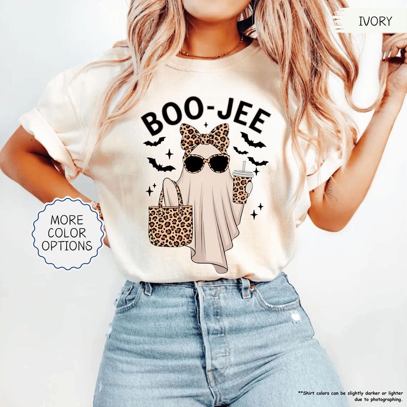 以下が含まれることがあります： 「BOO-JEE」の文字がプリントされたアイボリー色のTシャツ。サングラスとヒョウ柄のリボンをつけたお化けが、ヒョウ柄のトートバッグとコーヒーカップを持っています。黒いコウモリと星がお化けを囲んでいます。「MORE COLOR OPTIONS」は青い円の中にあります。