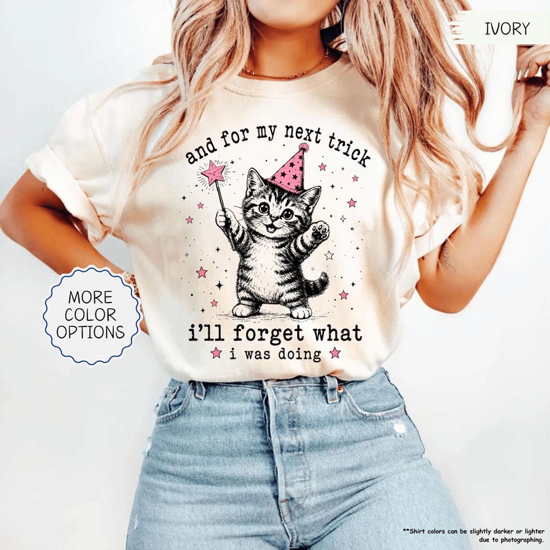 May include: An ivory t-shirt featuring a graphic of a kitten wearing a pink party hat and holding a star-tipped wand. The text reads, "and for my next trick i'll forget what i was doing," surrounded by small pink stars and black sparkles.