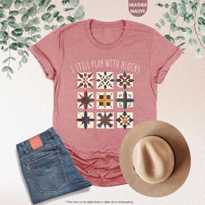 May include: A heather mauve t-shirt with the text "I Still Play With Blocks" and a pattern of colorful quilt blocks.