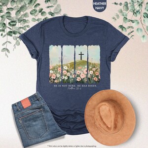 May include: A navy blue t-shirt with a watercolor design of a cross in a field of flowers. The text "HE IS NOT HERE. HE HAS RISEN. Matthew 28:6" is printed below the design.