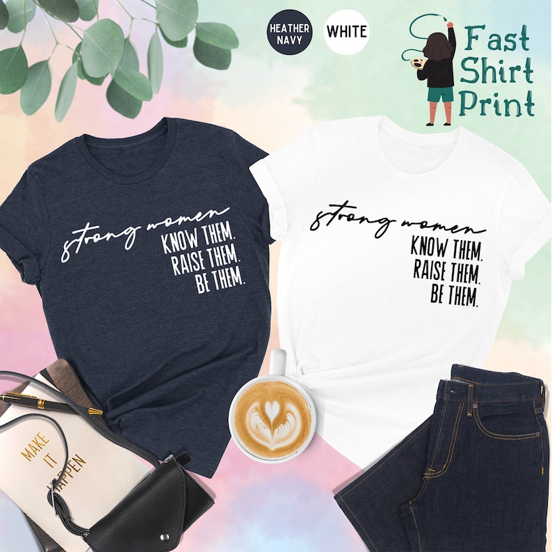 K&ouml;nnte beinhalten: Zwei T-Shirts mit dem Text "strong women know them. raise them. be them." in Schwarz auf einem wei&szlig;en T-Shirt und in Wei&szlig; auf einem helio-marinefarbenen T-Shirt.
