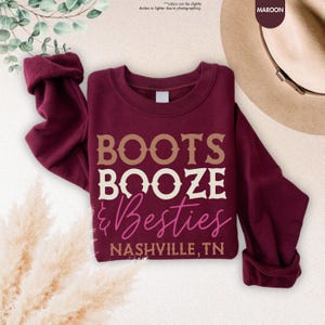 Pu&ograve; includere: Felpa bordeaux con la scritta "BOOTS BOOZE & Besties NASHVILLE, TN" in un design a strati. Le maniche sono arrotolate. Un cappello &egrave; visibile sullo sfondo. La felpa &egrave; piegata.