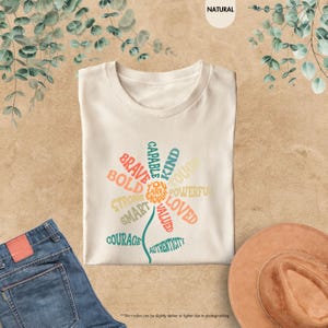 Pode incluir: Camiseta creme com um design floral composto por palavras positivas como "Brave", "Kind" e "Loved". A camiseta &eacute; combinada com jeans azuis e um chap&eacute;u marrom. O texto "You are enough" est&aacute; no centro da flor.