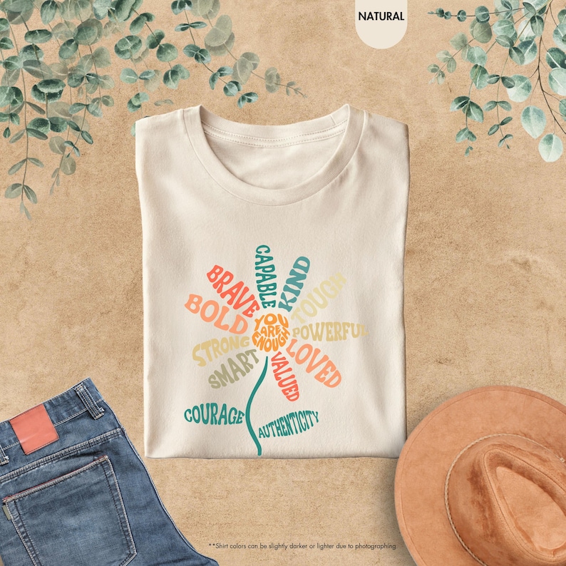 Pode incluir: Camiseta creme com um design floral composto por palavras positivas como "Brave", "Kind" e "Loved". A camiseta &eacute; combinada com jeans azuis e um chap&eacute;u marrom. O texto "You are enough" est&aacute; no centro da flor.
