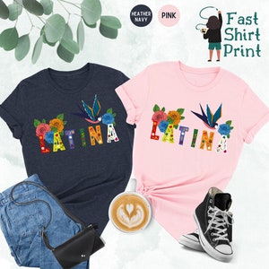 May include: Two t-shirts with the word "LATINA" printed in colorful letters with floral and bird designs. The shirts are available in heather navy and pink.