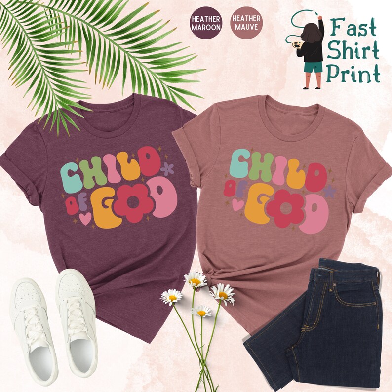 Op de afbeelding: Twee T-shirts met de tekst "Child of God" in een retro, kleurrijk lettertype. De shirts zijn heide-maron en heide-paars.