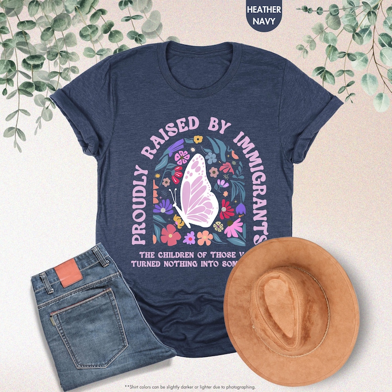 May include: A navy blue t-shirt with the text "PROUDLY RAISED BY IMMIGRANTS" and a butterfly graphic. The image also shows a pair of blue jeans and a brown hat.