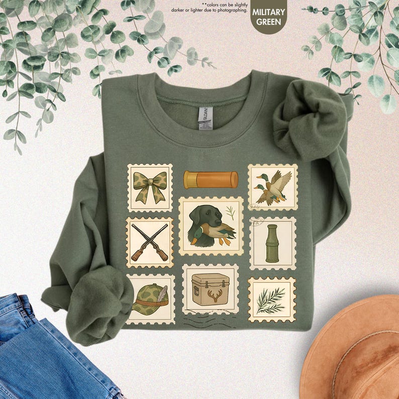 K&ouml;nnte beinhalten: Milit&auml;rgr&uuml;nes Sweatshirt mit Jagd-Postwertzeichen-Designs. Die Briefmarken zeigen Bilder von einem Bogen, Schrotpatronen, Enten, einem Hund mit Ente, gekreuzten Gewehren, einem Jagdhut, einer K&uuml;hlbox und einer Flasche.