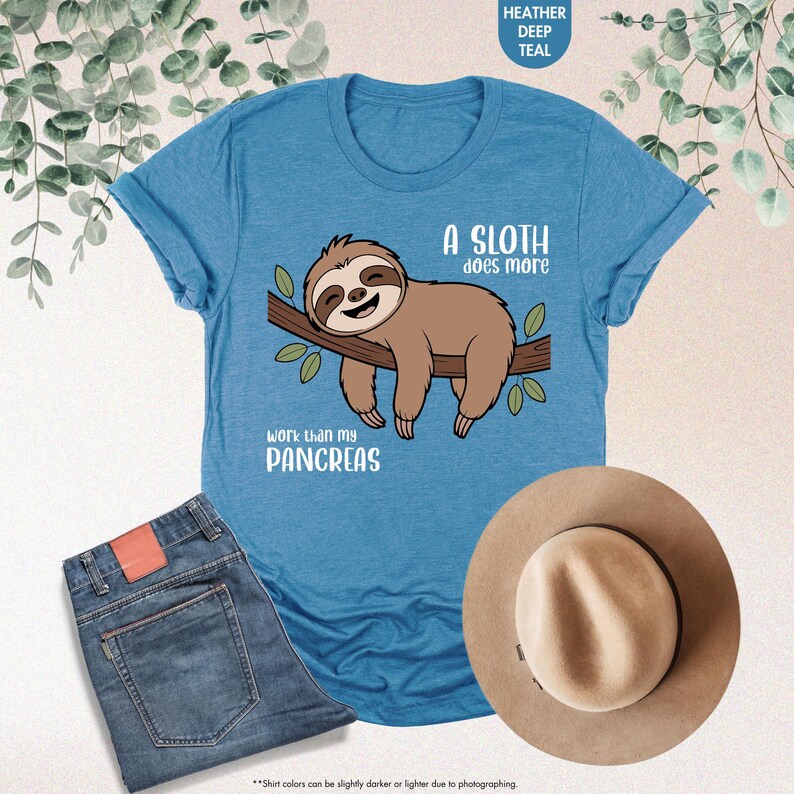 K&ouml;nnte beinhalten: Ein tief tealfarbenes T-Shirt mit einem Faultier-Motiv auf einem Ast und dem Text "A sloth does more work than my pancreas". Das Bild zeigt auch eine blaue Jeans und einen beigen Hut.