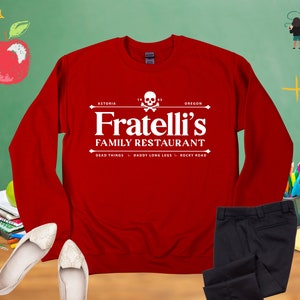 May include: Red long-sleeved sweatshirt with a white graphic of a skull and crossbones and the text "Fratelli's Family Restaurant" with the words "Astoria, Oregon" and "Dead Things, Daddy Long Legs, Rocky Road" underneath.