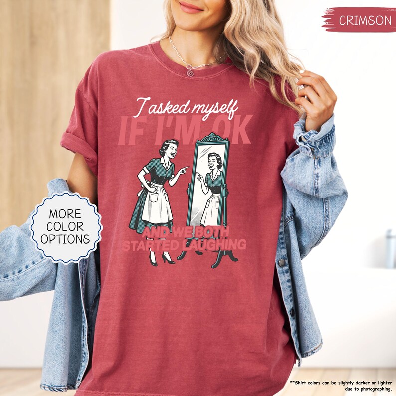May include: Crimson-colored t-shirt with a retro illustration of a woman looking in a mirror. The text reads "I asked myself IF I'M OK AND WE BOTH STARTED LAUGHING." The shirt has a vintage, washed look and is available in more colors.