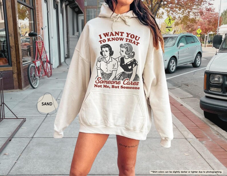 May include: Sand-colored hoodie with a drawstring hood and a front pocket. The graphic on the front features a vintage illustration of two women and the text "I WANT YOU TO KNOW THAT Someone Cares Not Me, But Someone."