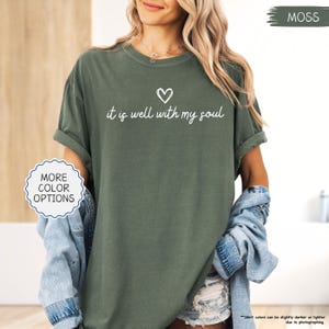 K&ouml;nnte beinhalten: Ein moosgr&uuml;nes T-Shirt mit hochgekrempelten &Auml;rmeln. Das Shirt hat den Schriftzug "it is well with my soul" in wei&szlig;er Schrift und ein kleines Herzsymbol. Die Worte "More Color Options" stehen in einem wei&szlig;en Kreis.