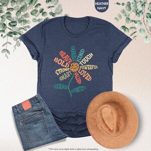 Pode incluir: T-shirt azul marinho mesclado com um design floral com palavras positivas como "corajoso" e "forte". A imagem tamb&eacute;m inclui jeans azuis e um chap&eacute;u marrom, sugerindo um traje casual.