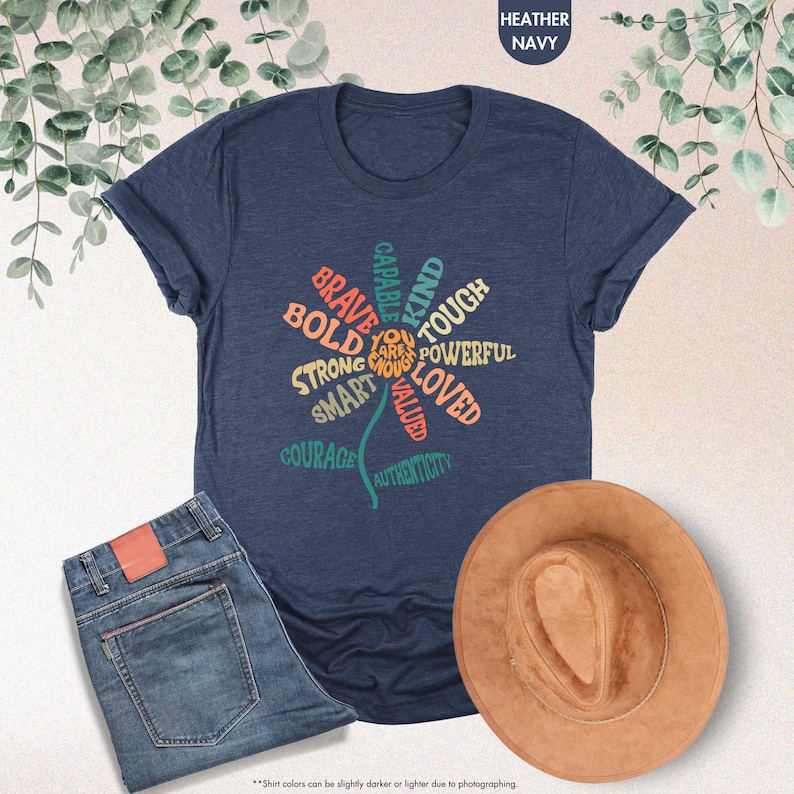 Pode incluir: T-shirt azul marinho mesclado com um design floral com palavras positivas como "corajoso" e "forte". A imagem tamb&eacute;m inclui jeans azuis e um chap&eacute;u marrom, sugerindo um traje casual.