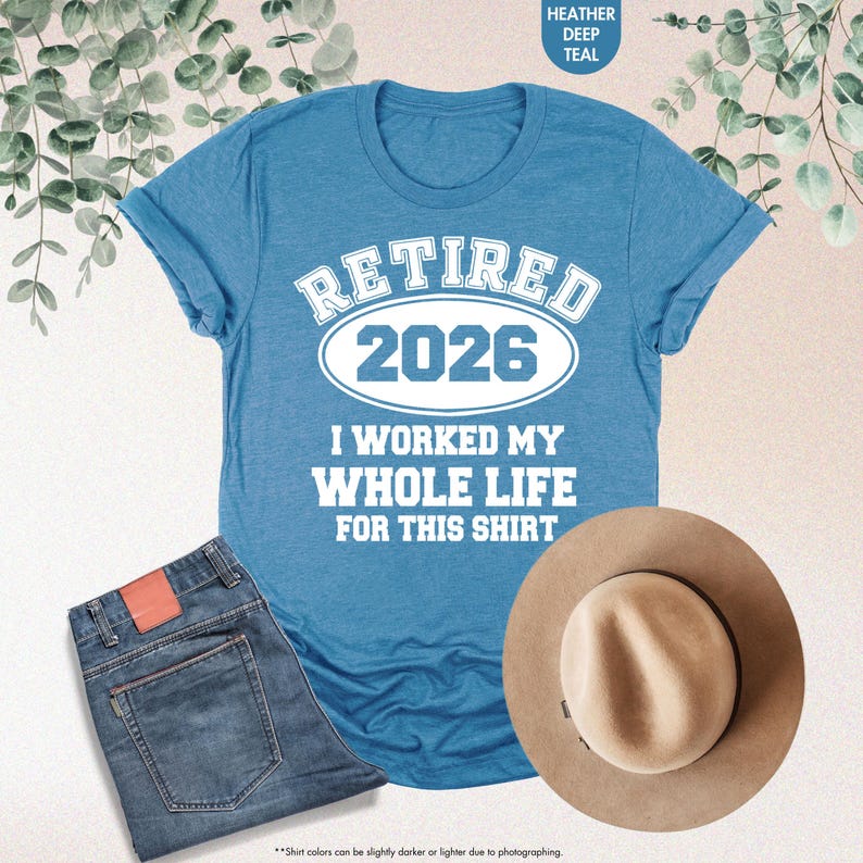 Puede incluir: Una camiseta de color verde azulado con el texto "RETIRED 2026 I WORKED MY WHOLE LIFE FOR THIS SHIRT". La camiseta se combina con vaqueros azules y un sombrero fedora color canela. La imagen tambi&eacute;n incluye una ramita de vegetaci&oacute;n.