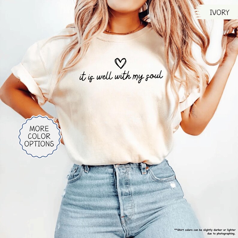 K&ouml;nnte beinhalten: Elfenbeinfarbenes T-Shirt mit dem Schriftzug "it is well with my soul" und einem Herzsymbol. Das Shirt hat hochgekrempelte &Auml;rmel und wird mit einer blauen Jeans kombiniert. Ein Knopf auf dem Shirt sagt "More Color Options".