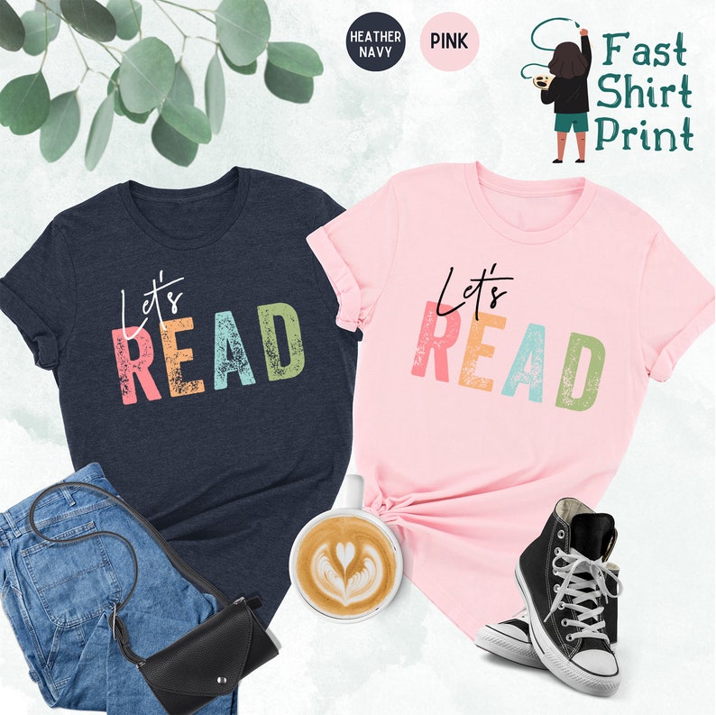 May include: Two t-shirts with the text "Let's Read" in colorful, distressed letters. The shirts are in heather navy and pink.