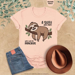 K&ouml;nnte beinhalten: Hellrosa T-Shirt mit einem braunen Faultier, das auf einem Ast mit gr&uuml;nen Bl&auml;ttern h&auml;ngt. Der Text lautet: "Ein Faultier arbeitet mehr als meine Bauchspeicheldr&uuml;se." Blaue Jeans und ein brauner Hut sind ebenfalls im Bild zu sehen.