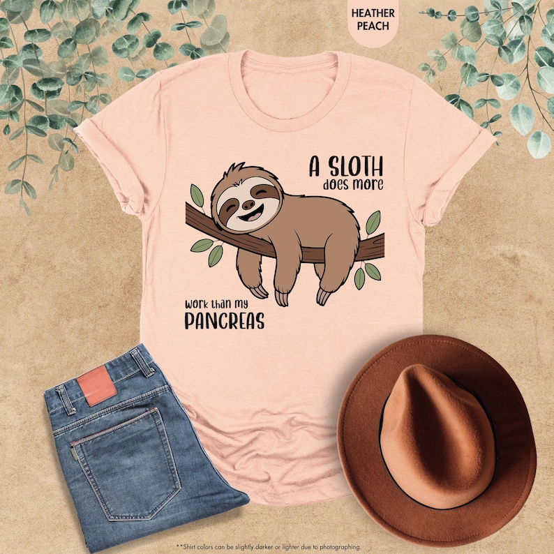 K&ouml;nnte beinhalten: Hellrosa T-Shirt mit einem braunen Faultier, das auf einem Ast mit gr&uuml;nen Bl&auml;ttern h&auml;ngt. Der Text lautet: "Ein Faultier arbeitet mehr als meine Bauchspeicheldr&uuml;se." Blaue Jeans und ein brauner Hut sind ebenfalls im Bild zu sehen.