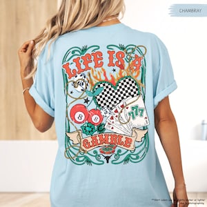 Puede incluir: Una camiseta azul claro con un dise&ntilde;o gr&aacute;fico de un coraz&oacute;n con un patr&oacute;n a cuadros, cartas de juego, una cuerda y una bola 8. El dise&ntilde;o gr&aacute;fico est&aacute; rodeado por un borde verde y dorado y el texto "Life is a Gamble".
