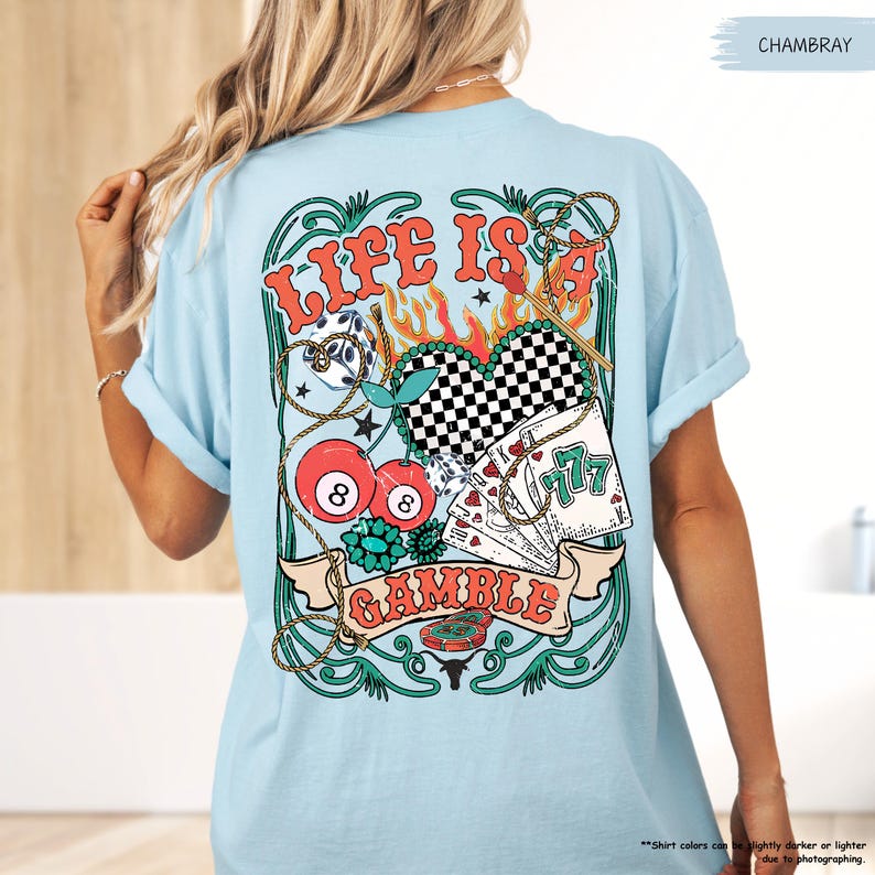 Puede incluir: Una camiseta azul claro con un dise&ntilde;o gr&aacute;fico de un coraz&oacute;n con un patr&oacute;n a cuadros, cartas de juego, una cuerda y una bola 8. El dise&ntilde;o gr&aacute;fico est&aacute; rodeado por un borde verde y dorado y el texto "Life is a Gamble".