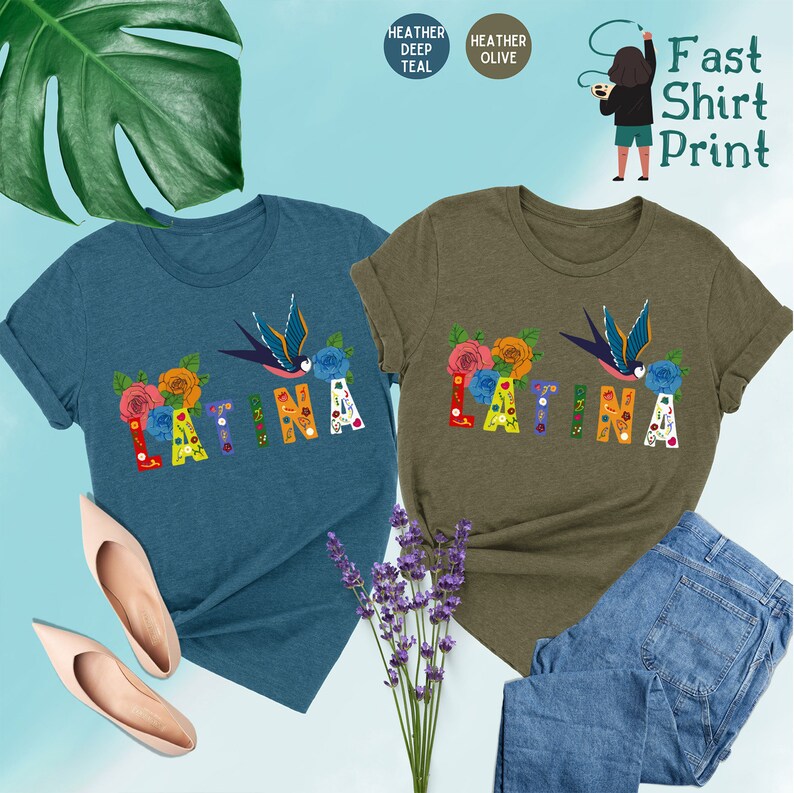May include: Two t-shirts with the word "LATINA" printed on them in colorful letters with floral and bird designs. The shirt on the left is a deep teal color and the shirt on the right is a heather olive color.