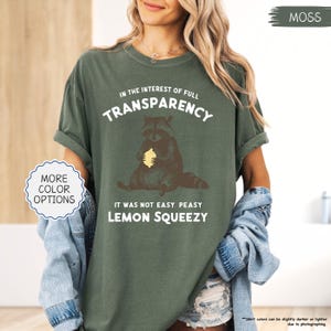 May include: Moss green t-shirt with the text "IN THE INTEREST OF FULL TRANSPARENCY" and "IT WAS NOT EASY PEASY LEMON SQUEEZY". The shirt features a raccoon graphic holding a lemon. "MORE COLOR OPTIONS" is in a white circle.