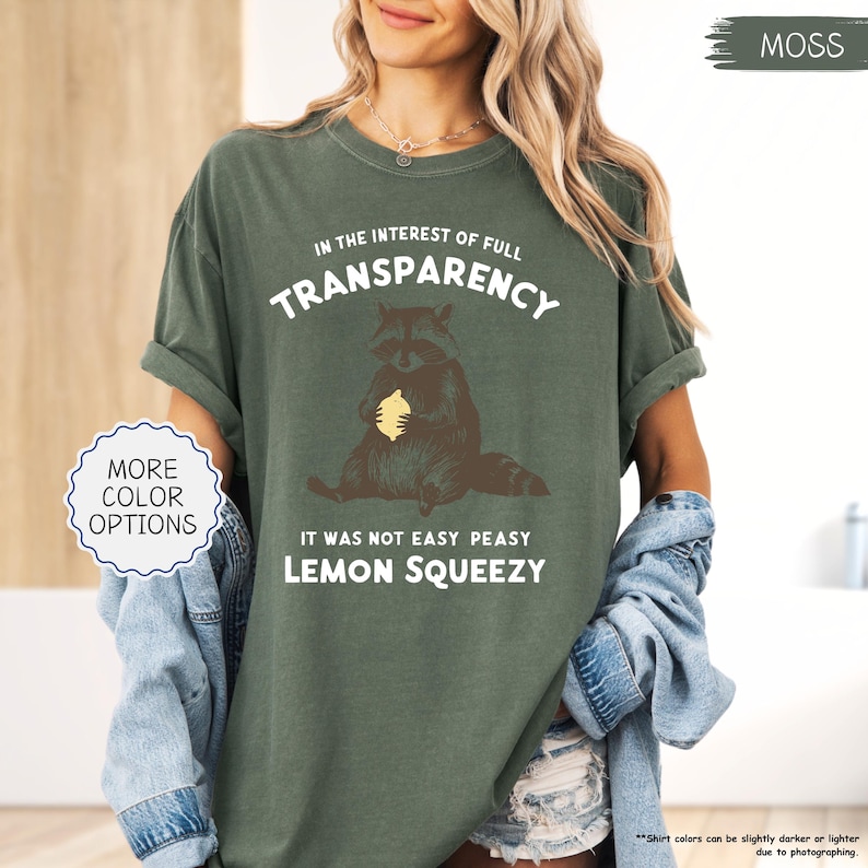May include: Moss green t-shirt with the text "IN THE INTEREST OF FULL TRANSPARENCY" and "IT WAS NOT EASY PEASY LEMON SQUEEZY". The shirt features a raccoon graphic holding a lemon. "MORE COLOR OPTIONS" is in a white circle.