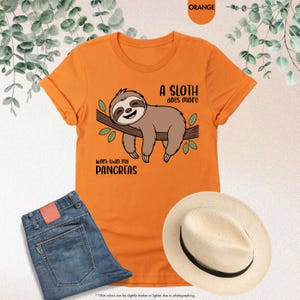 K&ouml;nnte beinhalten: Orangefarbenes T-Shirt mit einem Faultier-Motiv und dem Text "A SLOTH does more work than my PANCREAS." Das Bild zeigt auch eine blaue Jeans und einen Strohhut.