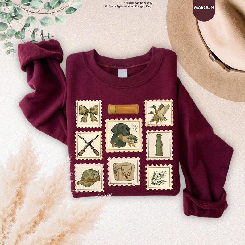 K&ouml;nnte beinhalten: Burgunderfarbenes Sweatshirt mit Jagd-Postwertzeichen-Designs. Die Briefmarken zeigen eine Schleife, ein Fernglas, Enten, gekreuzte Gewehre, einen Hund mit einer Ente, eine Flasche, einen Jagdhut, eine K&uuml;hlbox und Bl&auml;tter. Der Text "colors can be slightly darker or lighter due to photographing" ist sichtbar.