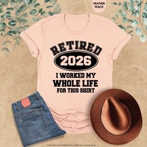 Puede incluir: Una camiseta color melocot&oacute;n con el texto "RETIRED 2026 I WORKED MY WHOLE LIFE FOR THIS SHIRT" en negro. La imagen tambi&eacute;n incluye unos vaqueros azules y un sombrero marr&oacute;n.