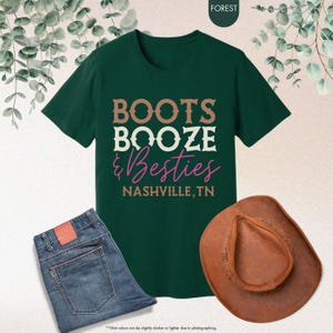Pu&ograve; includere: Una maglietta verde scuro con la scritta "BOOTS BOOZE & Besties NASHVILLE, TN" in marrone, bianco e rosa. L'immagine include anche un paio di jeans blu e un cappello da cowboy marrone, suggerendo un tema country.