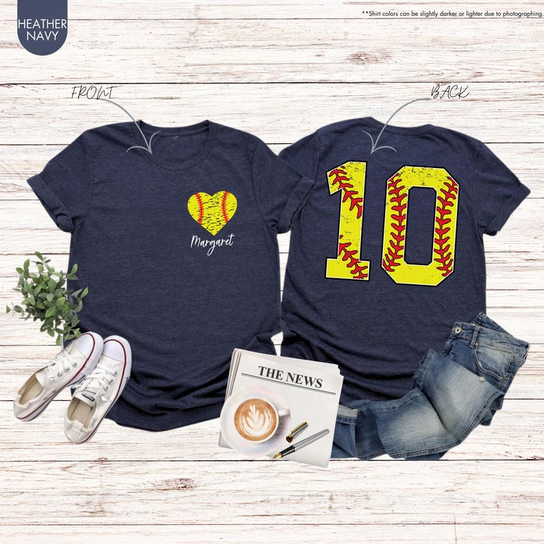 K&ouml;nnte beinhalten: Zwei marineblaue T-Shirts mit Softball-Motiv. Das vordere Shirt hat ein gelbes Softball-Herz mit roten N&auml;hten und den Namen "Margaret" darunter gedruckt. Das hintere Shirt hat eine gro&szlig;e gelbe Nummer "10" mit roten N&auml;hten, die einem Softball &auml;hnelt.